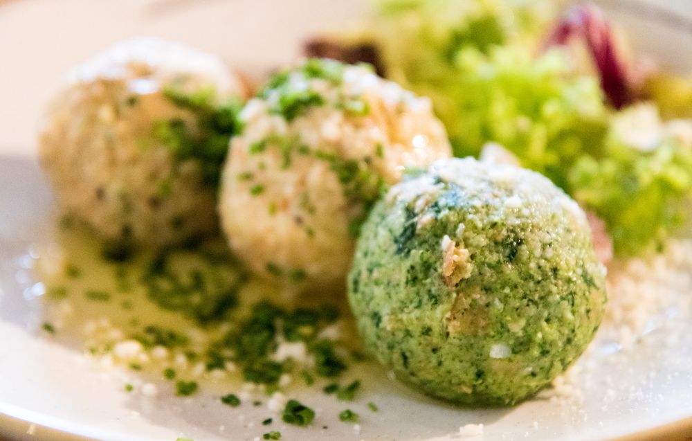 Teller mit drei Südtiroler Knödeln – darunter ein Spinatknödel und zwei Speck- bzw. Käseknödel – angerichtet in zerlassener Butter, bestreut mit Schnittlauch und Parmesan, dazu ein gemischter Blattsalat im Hintergrund.