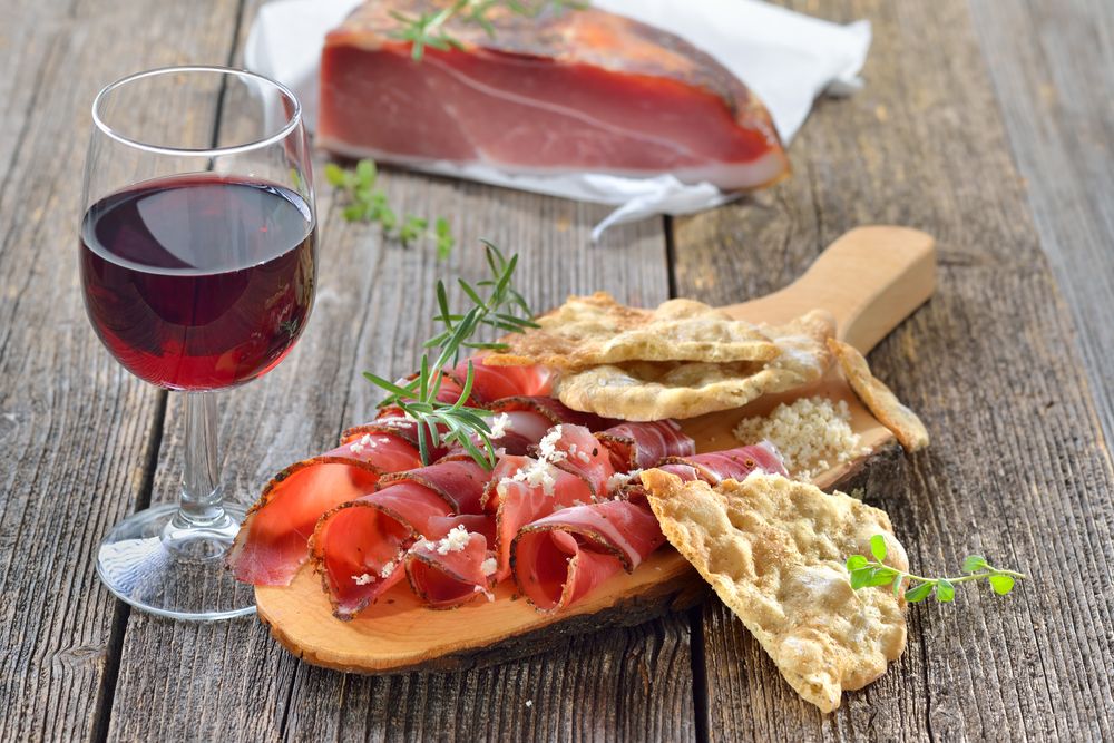 Holzbrett mit hauchdünn geschnittenem Südtiroler Speck, dekoriert mit frischem Rosmarin, knusprigem Schüttelbrot und einem Glas Rotwein auf rustikalem Holztisch – im Hintergrund ein Stück ungeschnittener Speck.
