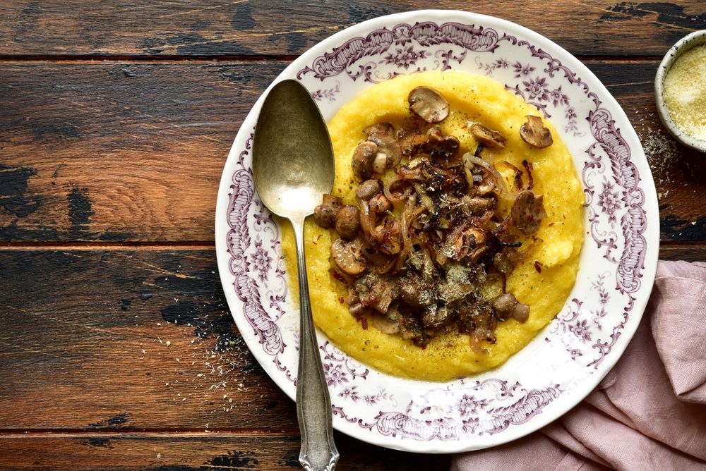 Traditionelle Polenta mit Pilzragout und Zwiebeln auf verziertem Porzellanteller, angerichtet auf rustikalem Holztisch mit Löffel und Parmesanschale