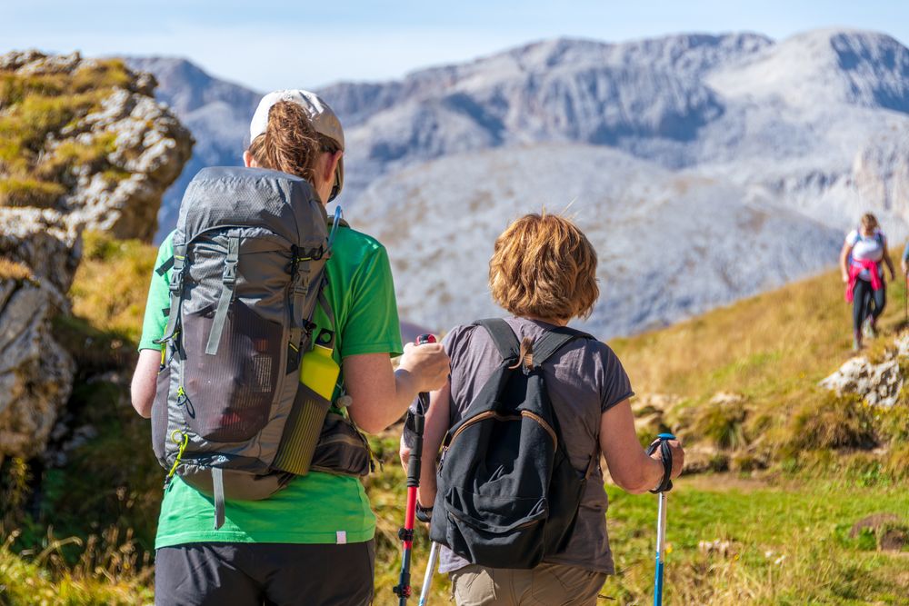 „Zwei Wanderer mit Rucksäcken und Wanderstöcken von hinten auf einem sonnigen Bergpfad in den Dolomiten in Südtirol – im Hintergrund beeindruckende Felsgipfel und eine weitere Wanderin auf dem Weg.