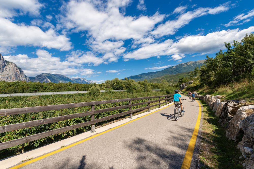 Radfahrer auf einem gut ausgebauten Radweg mit gelber Markierung entlang eines Sees im Trentino – umgeben von üppigem Grün, Bergen und einem teilweise bewölkten blauen Himmel