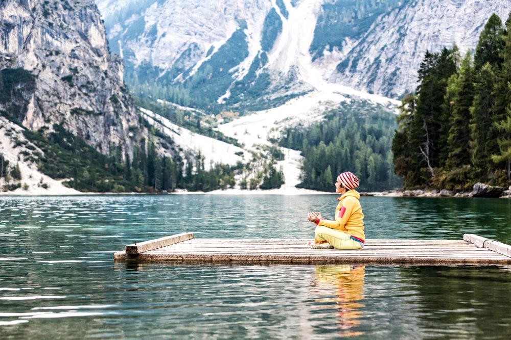 Person in bunter Kleidung bei einer Yoga-Übung im Schneidersitz auf einem Holzsteg am Pragser Wildsee im Pustertal – umgeben von türkisfarbenem Wasser, dichten Wäldern und steil aufragenden Dolomitengipfeln